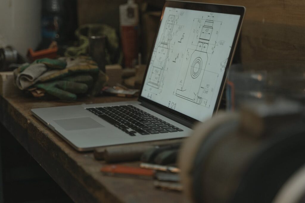 Laptop showing a technical drawing, surrounded by tools and gloves in a workshop setting.